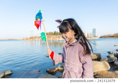 a happy little girl stands on a riverside rock, holding a colorful pinwheel. a happy little girl stands on a riverside rock, holding a colorful pinwheel. 141312079