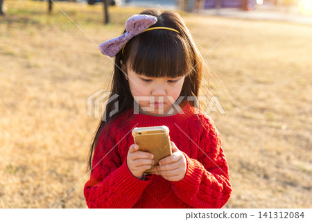 a little girl in a red sweater looks down and uses a smartphone in the grass. a little girl in a red sweater looks down and uses a smartphone in the grass. 141312084