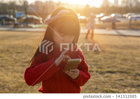 a little girl in a red sweater looks down and uses a smartphone in the grass. 141312085