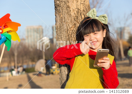a smiling girl leans against a tree trunk and gives a thumbs up while holding a smartphone. a smiling girl leans against a tree trunk and gives a thumbs up while holding a smartphone. 141312089