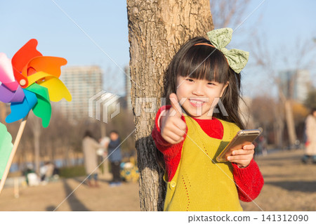 a smiling girl leans against a tree trunk and gives a thumbs up while holding a smartphone. 141312090