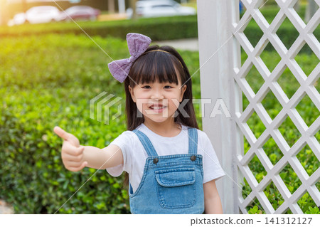 A joyful young girl with a bow stands on a park path, raising one arm up high with a bright smile. A joyful young girl with a bow stands on a park path, raising one arm up high with a bright smile. 141312127