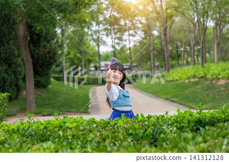 A joyful young girl with a bow stands on a park path, raising one arm up high with a bright smile. A joyful young girl with a bow stands on a park path, raising one arm up high with a bright smile. 141312128