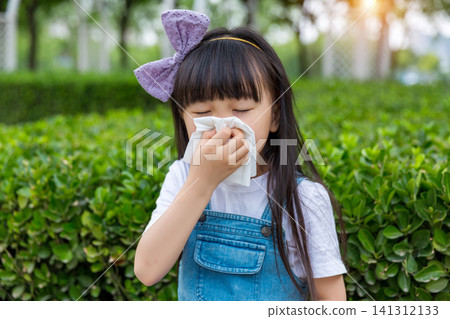 A young girl with a bow headband uses a white tissue to blow her nose while standing in a park with hedges. A young girl with a bow headband uses a white tissue to blow her nose while standing in a park with hedges. 141312133