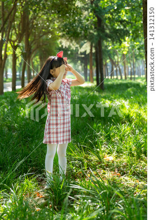 A joyful young girl in a plaid dress stands in a lush green grassy field, using both hands to make an eye-glass gesture. A joyful young girl in a plaid dress stands in a lush green grassy field, using both hands to make an eye-glass gesture. 141312150