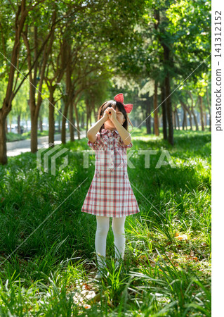 A joyful young girl in a plaid dress stands in a lush green grassy field, using both hands to make an eye-glass gesture. A joyful young girl in a plaid dress stands in a lush green grassy field, using both hands to make an eye-glass gesture. 141312152