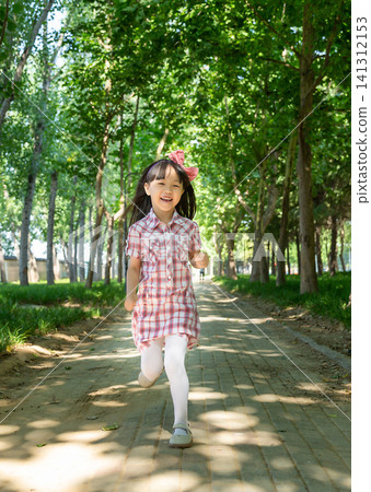 Cheerful little girl running forward on a brick path in a lush green park during summertime. 141312153