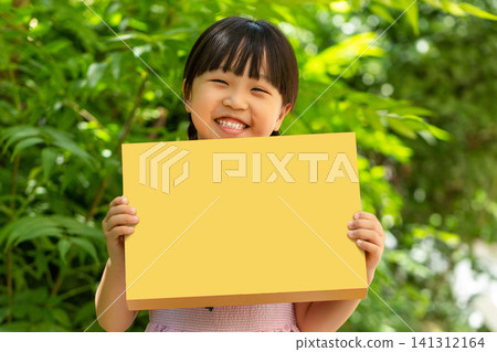 Cheerful child holding a blank yellow cardboard rectangle sign against a vibrant green garden backdrop. Cheerful child holding a blank yellow cardboard rectangle sign against a vibrant green garden backdrop. 141312164