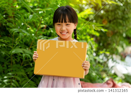 Cheerful child holding a blank yellow cardboard rectangle sign against a vibrant green garden backdrop. 141312165