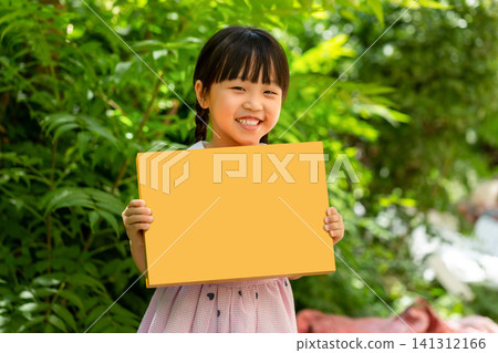 Cheerful child holding a blank yellow cardboard rectangle sign against a vibrant green garden backdrop. 141312166