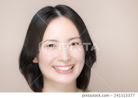 一位美麗的日本女子，背景是粉米色，笑容甜美，展現出她無瑕的肌膚和自然的妝容，直視著鏡頭。 141313377