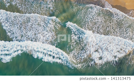 Horizontal top down aerial view of rolling turquoise sea waves with white foam breaking on a sandy shore creating a beautiful natural coastal background 141313475