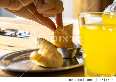 A hand dipping a golden cheese bread into a small metallic bowl on a stainless tray, showcasing a traditional Brazilian snack experience for culinary blogging or restaurant menu use A hand dipping a golden cheese bread into a small metallic bowl on a stainless tray, showcasing a traditional Brazilian snack experience for culinary blogging or restaurant menu use 141313588
