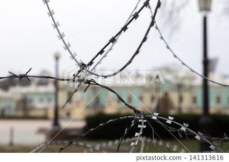 A sharp razor wire fence stands against a blurred historical building background, highlighting themes of restricted access, strict security, defense, and urban social isolation barriers 141313616