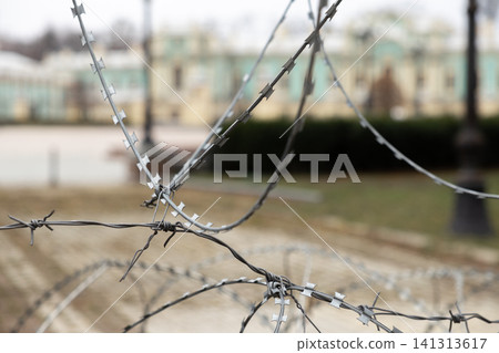 Sharp coiled razor wire creates a physical barrier before an ornate historical building, symbolizing strict restriction, protection, geopolitical conflict, or deep civic tension today Sharp coiled razor wire creates a physical barrier before an ornate historical building, symbolizing strict restriction, protection, geopolitical conflict, or deep civic tension today 141313617