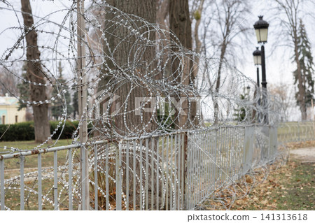 A sharp razor wire coil rests atop a metallic perimeter fence in a public park, conveying restricted access and high security measures within an urban environment during wintertime A sharp razor wire coil rests atop a metallic perimeter fence in a public park, conveying restricted access and high security measures within an urban environment during wintertime 141313618