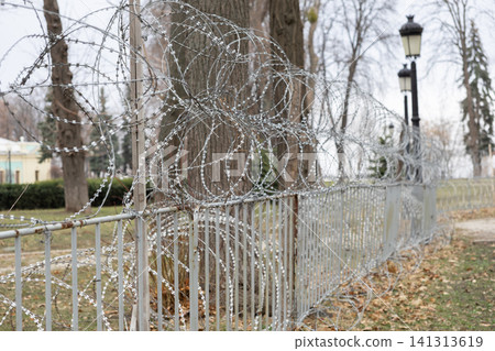 Coils of razor wire atop a metal park fence, creating a barrier in a public outdoor space, symbolizing strict security, restricted access, and modern urban danger or fortification Coils of razor wire atop a metal park fence, creating a barrier in a public outdoor space, symbolizing strict security, restricted access, and modern urban danger or fortification 141313619