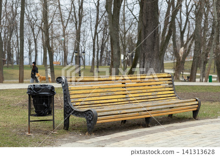 A rustic weathered yellow wooden bench sits by a black metal trash bin in a peaceful park during early spring, evoking a sense of calm reflection in an urban natural landscape area A rustic weathered yellow wooden bench sits by a black metal trash bin in a peaceful park during early spring, evoking a sense of calm reflection in an urban natural landscape area 141313628