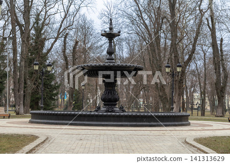 An ornate cast iron fountain stands in a quiet public city park during autumn, surrounded by bare trees under a cloudy sky to evoke a serene historical urban landscape for local travel 141313629