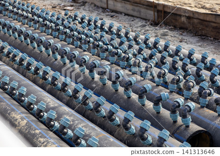 A series of black plastic irrigation pipes with light blue fittings aligned on a construction site, illustrating modern agricultural water management and efficient field infrastructure A series of black plastic irrigation pipes with light blue fittings aligned on a construction site, illustrating modern agricultural water management and efficient field infrastructure 141313646