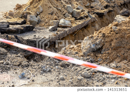 A deep trench excavated in an asphalt road for utility pipe repairs, surrounded by heaps of dirt and rocks, marked by red and white safety tape to warn pedestrians of construction A deep trench excavated in an asphalt road for utility pipe repairs, surrounded by heaps of dirt and rocks, marked by red and white safety tape to warn pedestrians of construction 141313651