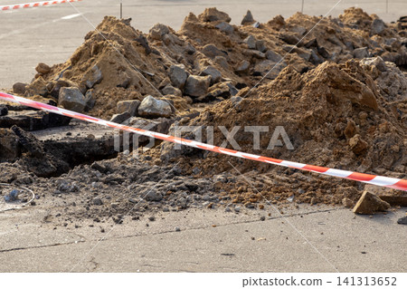 Road construction trench with piles of dug soil and rubble cordoned off by striped red white warning tape, marking an urban utility repair project site for infrastructure maintenance Road construction trench with piles of dug soil and rubble cordoned off by striped red white warning tape, marking an urban utility repair project site for infrastructure maintenance 141313652