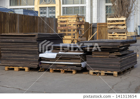Stacked plywood boards on wooden pallets sit on an asphalt lot near a building exterior, representing industrial storage, construction materials management, and warehouse logistics 141313654
