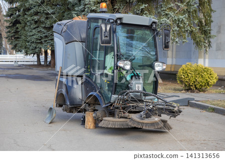 A small street sweeping machine sits stationary on wooden blocks during repair in a park, representing maintenance and industrial service work for clean urban public environments A small street sweeping machine sits stationary on wooden blocks during repair in a park, representing maintenance and industrial service work for clean urban public environments 141313656
