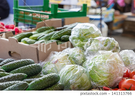Fresh green cucumbers and wrapped lettuce heads displayed in a cardboard box at a busy outdoor farmers market, showcasing raw seasonal vegetables for healthy eating and local grocery Fresh green cucumbers and wrapped lettuce heads displayed in a cardboard box at a busy outdoor farmers market, showcasing raw seasonal vegetables for healthy eating and local grocery 141313766