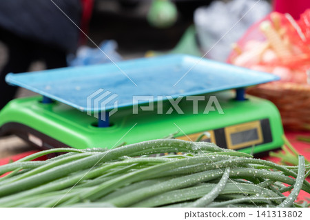 Fresh green spring onion stalks lie near electronic weighing scales on a red market table, highlighting natural food quality and local agriculture trade in an authentic outdoor 141313802