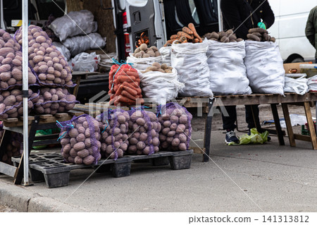 Fresh vegetables displayed in mesh bags and white sacks on wooden pallets at an outdoor farmers market stand, showcasing bulk local produce for sale in a rural grocery setting Fresh vegetables displayed in mesh bags and white sacks on wooden pallets at an outdoor farmers market stand, showcasing bulk local produce for sale in a rural grocery setting 141313812