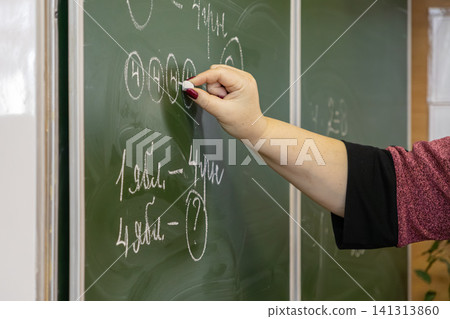 A dedicated teacher writes complex math exercises with white chalk on a dark green chalkboard in an elementary school classroom to foster academic learning for young pupils today A dedicated teacher writes complex math exercises with white chalk on a dark green chalkboard in an elementary school classroom to foster academic learning for young pupils today 141313860