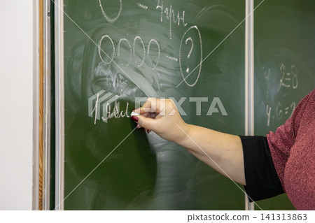 A teacher writes complex math exercises with white chalk on a green classroom blackboard to represent primary school education, academic instruction, and learning in a school A teacher writes complex math exercises with white chalk on a green classroom blackboard to represent primary school education, academic instruction, and learning in a school 141313863