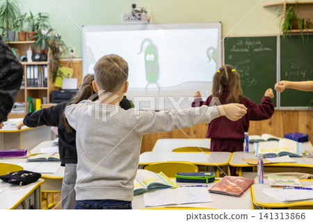 Primary school students perform active physical exercise breaks in a classroom while following a cartoon animation on a digital screen to maintain focus during daily lesson plans Primary school students perform active physical exercise breaks in a classroom while following a cartoon animation on a digital screen to maintain focus during daily lesson plans 141313866