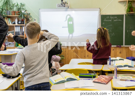 Schoolchildren stand and follow physical exercise while looking at a cartoon character on a digital screen during an interactive primary school lesson in a bright modern classroom Schoolchildren stand and follow physical exercise while looking at a cartoon character on a digital screen during an interactive primary school lesson in a bright modern classroom 141313867