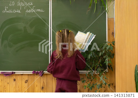 A young primary school student stands by a green chalkboard reading a textbook in a classroom, showcasing academic focus and learning engagement within a traditional school setting 141313871