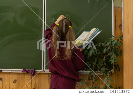A young schoolchild writes on a green chalkboard while holding a textbook in a classroom, representing primary education, academic growth, learning, concentration and development 141313872