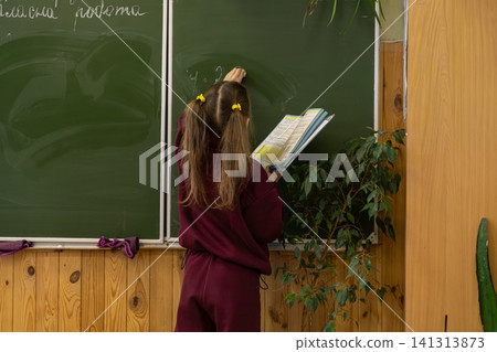 A young schoolchild writes on a green chalkboard in an elementary classroom, holding a textbook while studying during a lesson to represent academic focus in primary education A young schoolchild writes on a green chalkboard in an elementary classroom, holding a textbook while studying during a lesson to represent academic focus in primary education 141313873