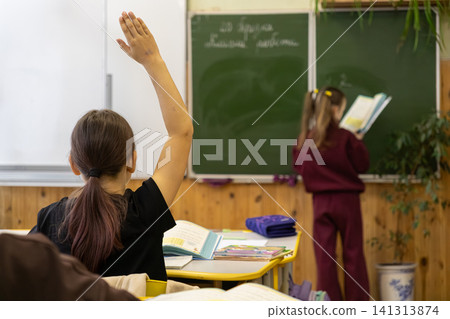 Schoolchild raising hand to ask question in primary school classroom with classmate studying at chalkboard, representing active student engagement and academic learning environment Schoolchild raising hand to ask question in primary school classroom with classmate studying at chalkboard, representing active student engagement and academic learning environment 141313874