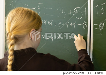 Schoolchild writing math equations with chalk on a green chalkboard in a primary school classroom, illustrating an active learning moment for young students in an academic setting 141313891