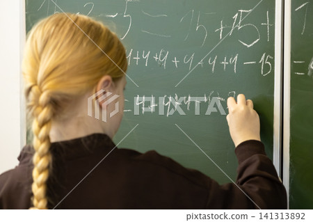 A young schoolchild writes complex math equations with white chalk on a green classroom board, representing focused learning and education for students developing academic skills 141313892
