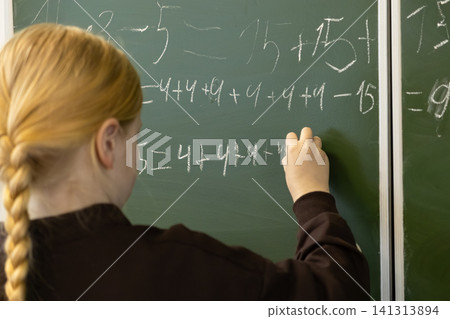 A young schoolchild writes complex math equations on a green classroom blackboard using white chalk while engaged in focused learning during an elementary school mathematics lesson A young schoolchild writes complex math equations on a green classroom blackboard using white chalk while engaged in focused learning during an elementary school mathematics lesson 141313894
