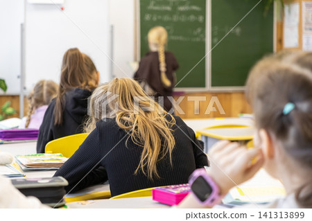 Students sit at desks in a bright primary school classroom while a classmate stands at the chalkboard to study math during an engaging academic lesson in a modern educational space Students sit at desks in a bright primary school classroom while a classmate stands at the chalkboard to study math during an engaging academic lesson in a modern educational space 141313899