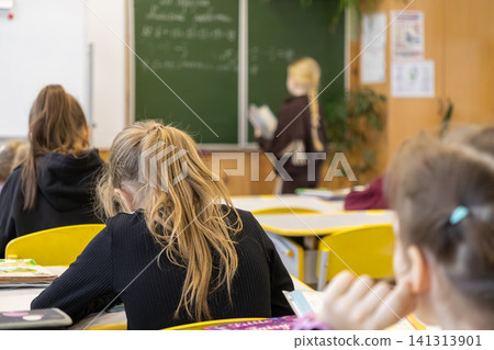 Students sitting at desks in a primary school classroom while a classmate stands at the chalkboard to present, reflecting an active learning environment during a daily lesson Students sitting at desks in a primary school classroom while a classmate stands at the chalkboard to present, reflecting an active learning environment during a daily lesson 141313901