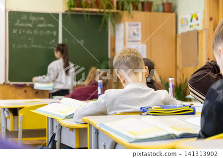 Schoolchildren study at wooden desks in an elementary classroom while a classmate writes on the green blackboard during an engaging math lesson focused on academic growth today 141313902