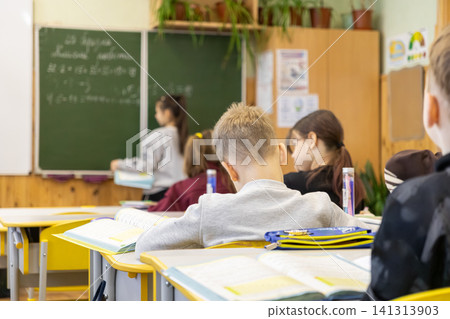 Schoolchildren sitting at wooden desks in an elementary classroom, watching their peer write on a green chalkboard during a math lesson, depicting active learning and education mood Schoolchildren sitting at wooden desks in an elementary classroom, watching their peer write on a green chalkboard during a math lesson, depicting active learning and education mood 141313903