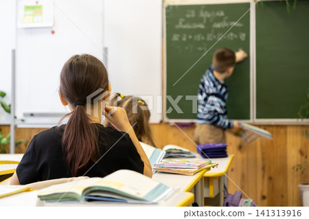 Young students sit at wooden desks in an elementary school classroom while a classmate writes on a green chalkboard to capture the focused atmosphere of primary education lessons Young students sit at wooden desks in an elementary school classroom while a classmate writes on a green chalkboard to capture the focused atmosphere of primary education lessons 141313916