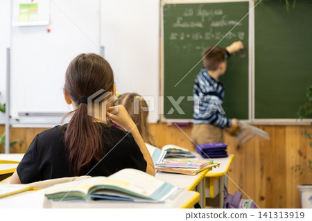 Students sit at wooden desks in a bright primary school classroom while a classmate writes on a green chalkboard, capturing a moment of academic engagement during a daily lesson Students sit at wooden desks in a bright primary school classroom while a classmate writes on a green chalkboard, capturing a moment of academic engagement during a daily lesson 141313919
