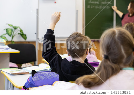 A young schoolchild raises their hand in a primary classroom during a lesson while the teacher writes on the green board, showing active participation and engagement in education A young schoolchild raises their hand in a primary classroom during a lesson while the teacher writes on the green board, showing active participation and engagement in education 141313921