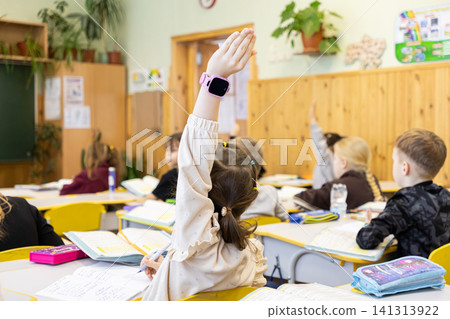 Schoolchildren sit at desks in an elementary classroom with raised hands to participate in a lesson, showing focus and engagement in a vibrant primary education learning environment Schoolchildren sit at desks in an elementary classroom with raised hands to participate in a lesson, showing focus and engagement in a vibrant primary education learning environment 141313922